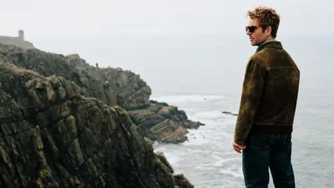 UKTV A man looking out to the side with the sea in front of him and cliff paths to the left.