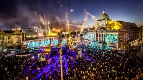 A night time arts event in Queen Victoria Square in Hull during the City of Culture in 2017. A large crowd watch as images are projected onto the City Hall and Ferens Art Gallery 