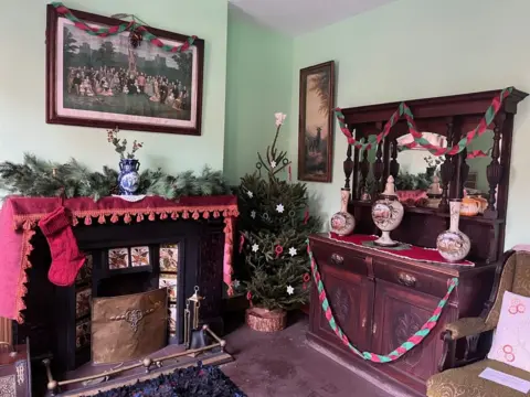 Christmas decorations are on the sideboard on the right and over a picture on the left that is hanging above the fireplace. A Christmas tree and another picture on the wall is in the middle.