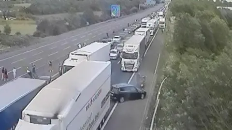 A motorway camera view showing a queue on one carriageway, People can be seen out of their cars. 