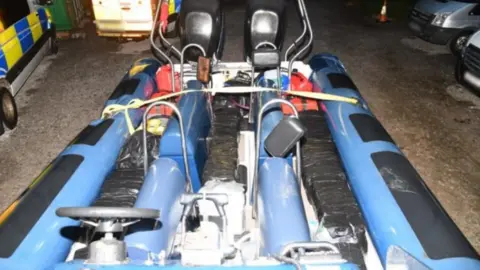 National Crime Agency A blue speedboat sits on the tarmac. Square parcels wrapped tightly in black binbags are stashed between the seats. Red fuel cans sit at the rear of the boat. Police vehicles are visible to the left of the image. 
