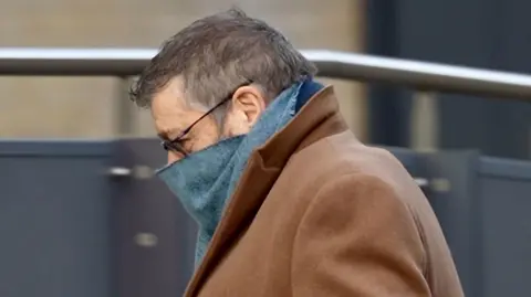 Forbes Scott arriving at court. He has short greying hair, glasses, a scarf pulled up to his nose and a brown coat