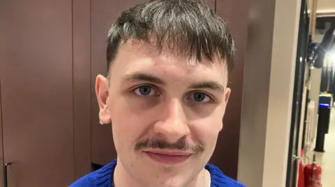 A young man, Turlough O'Hagan pictured wearing a blue jumper. He has brown hair and a dark moustache.