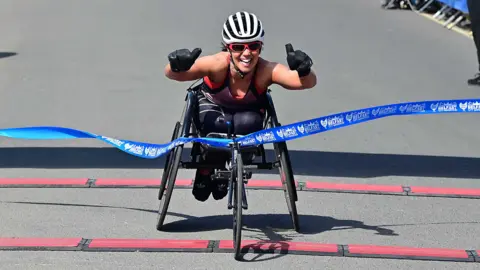 A lady in a wheelchair with a white helmet puts her thumbs up as she crosses through the blue ribbon. She is wearing red glasses and is wearing black gloves. She is wearing a vest with leggings.