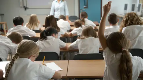 Children wearing white shirts sitting in a classroom. One child has her hand up. The photo is taken from behind the children so their faces are not visible.