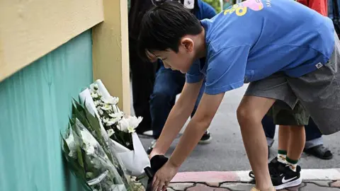 AFP via Getty Images A boy in a blue t-shirt and shorts lays a bouquet of white flowers on the ground. 