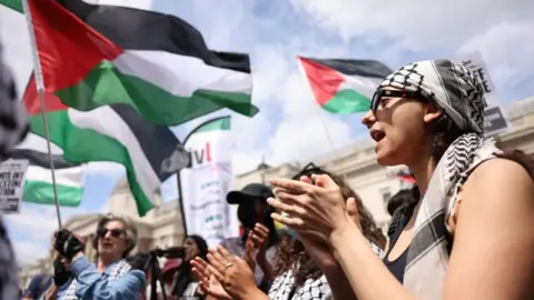 A woman from side on clapping and shouting, as people behind her hold Palestinian flags