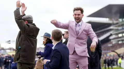 PA Media Racegoers are cheering during a race. On the left is a man in a dark green Barbour jacket clapping with his hands above his head. In the centre is a man in a dark blue blazer looking up at a man who is facing the camera, cheering, in a full lilac suit. He is holding the top of a pint cup.