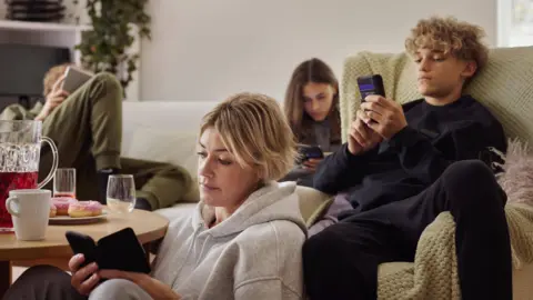 Getty Images A family (mother, two children, father) sitting down in the living room - on a sofa and the floor - all looking at their phones separately and looking unimpressed.