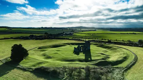 An aerial image of church ruins sitting among earthworks in a green landscape with blue cloudy skies