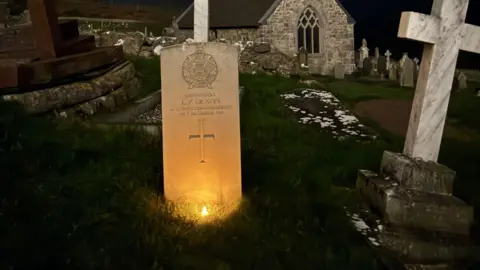 Home Front Museum, Llandudno A white Portland stone war grave stands in the centre of the shot.  It's night time and it is lit up by the warm orange glow of a battery night light candle.  In the background are other graves, and the arched window of a small church.