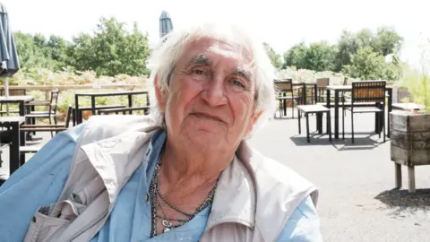 John Fairhall/BBC A man with messy white hair and wrinkled facial features sits outdoors at an open-air cafe terrace with wooden tables and chairs. He wears a light blue collared shirt, multiple silver chain necklaces, and a light-colored outdoor vest.