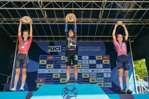 Red On Sports/Angus MacKinnon Three women on a podium hold their trophies in the air