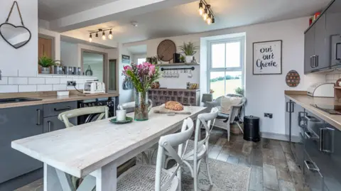 Michael Speak A modern grey and creme coloured kitchen with a large dining table and four chairs in the middle and cipboards and work surfaces on either side. 