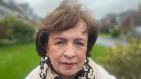Diane Dodds has shoulder length brown hair, is wearing makeup with pink lipstick. She is wearing a cream jumper and a black and cream leopard print scarf.