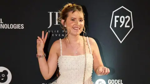 Issie is at the Champions for Children gala dinner. She is posing for the camera with her right hand making a peace sign and she is smiling. She is wearing a white dress embellished with pearls