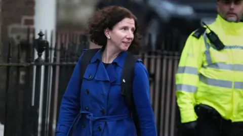 Anneliese Dodds is seen leaving a cabinet meeting, she is wearing a blue coat and backpack. A security officer in high vis is in the background. 