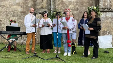 Malmesbury's Ukrainian choir wearing traditional clothing and singing into microphones in front of the Abbey wall.