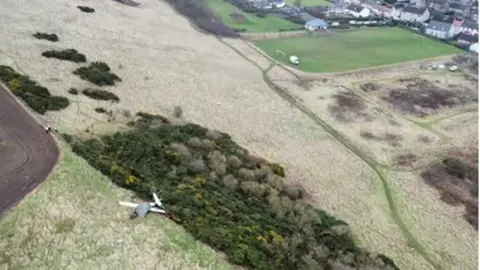 Air Accident Investigation Branch An aerial photgraph shows a green tarpaulin covering the wreckage of a small plane. Some white metallic parts protrude from under the cover. The plane is on a grassy field next to an area of trees and bushes. Houses can be seen beyond the fields and a playing field with goalposts.