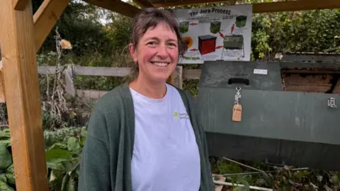 A compost mentor from Sustainable South Hams, Melissa Harvey, standing in front of a 'hot compost' tumbling machine. She is wearing a tee shirt and green cardigan.  The tumbling machine is dark green and there are two doors on the top, one of them is open.   