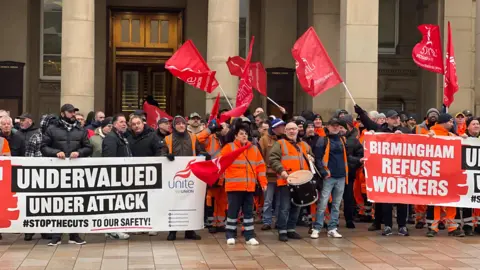 A group of people, some in hi vis clothing, stand outside the columns of a building. They are flying red flags and holding signs reading: "Undervalued. Under attack. hashtag stop the cuts to our safety".