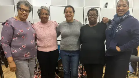 BBC A partir da esquerda, Beverley Hepburn-Henderson, Kathy Hepburn-Hinds, Evadne Anderson, Avril Bartholomew e Angela Brown-Johnson estão todas abraçadas. Beverley está com um suéter roxo e calças e óculos cinza. Kathy está vestindo um suéter rosa e calça preta e está de óculos. Evadne está com blusa cinza e jeans. Avril está vestindo blusa e calça pretas e óculos e Angela está com um moletom azul com lenço azul e calça escura.