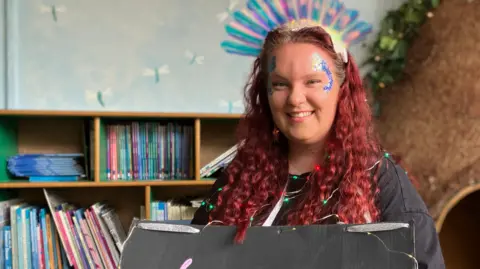Teacher Lauren Noone with long red, curly hair and glittery make up. She is a holding a sign representing her word 'luminescent'