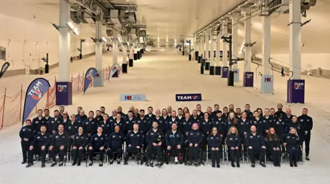 Team UK sit for the camera. The team are dressed in a navy blue uniform. They appear to be at the bottom of a slope at an indoor ski slope.