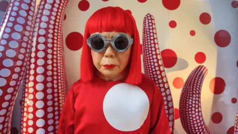 Getty Images Yayoi Kusama with red wig, red top with a large giant spot and goggles, in front of a red and white spotty tentacle-like artwork