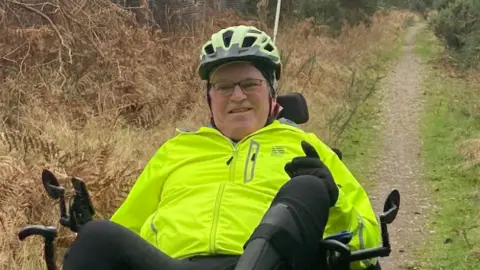 Supplied Nick Apperley wearing a hi-vis jacket and black trousers and a helmet while sitting and cycling on a trike on a woodland trail. 