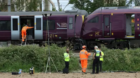 PA Media Two trains that have collided into each other. There are people wearing hi-vis jackets in the image. One person wearing orange hi-vis is walking on to the train and the other three are standing next to the train tracks.