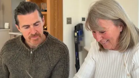 On the left is a middle aged man, he has brown grayish hair and is wearing a brown quarter zip - he has a mustache and is looking down. To his right is a woman with short grayish blonde hair who is smiling and looking down 