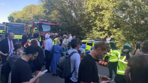 A crowd of people stand outside near police, fire brigade and ambulance vehicles. A number of people in yellow jackets and green helmets can also be seen in the image.