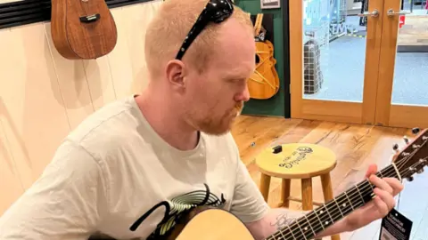 Craig Brown Craig Brown, seated, playing the guitar in a shop with guitars mounted on the wall behind him