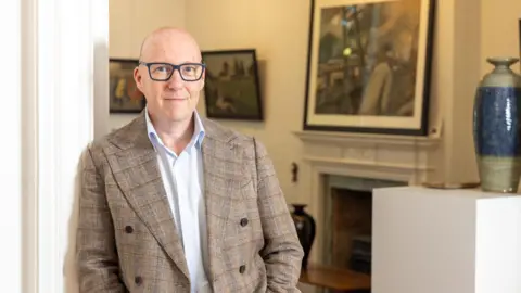 Steve Swallow. He is bald and has blue eyes. He wears glasses with a blue frame, a white shirt and chequered beige and brown jacket. He is leaning on a wall with artworks in the background. 