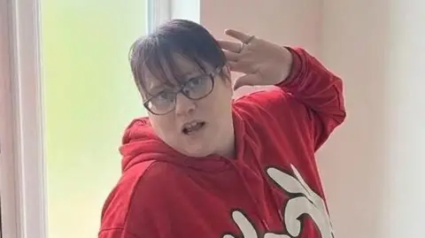 Alison Thompson has one arm bent behind her head as she poses for the camera. She is standing in front of a glass door. She has short dark hair, wears dark framed glasses and is wearing a red jumper with white writing.