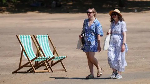 PA Media Two women walk infront of two deckchairs. The ground is yellow and parched.