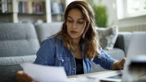 A woman with long dark hair and wearing a blue denim jacket over a black top looks down at a piece of paper while sat at a desk, in front of a grey sofa, with an open laptop in front of her