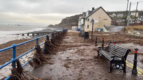 Weather Watcher Stephen Wright a seafront walk, with typical seafront buildings and benches, but the railings are draped with seaweed and the path and car park are covered in seaweed which has been blown up from the beach as the result of stormy weather. The sky is grey and the sea is grey and choppy.