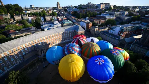 A group of 11 colourful hot air balloons are seen from above. They are fully inflated on College Green in Bristol as a publicity event for the 2026 Bristol International Balloon Fiesta. In the background Park Street is visible leading uphill towards the Wills Memorial Building, a large stone tower, in the distance