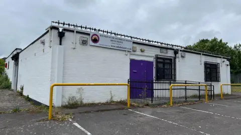 The single-storey brick building has been painted white. It has a purple door and bars on the windows. It stands in a car park and has barbed wire on the roof.