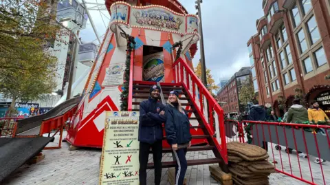 BBC/Kaya Black Two workers posing below a brightly decorated read and white helter-skelter ride.