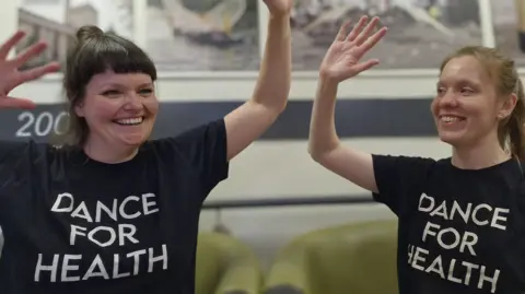 Dance for Health Two women, in matching T-shirts, with their arms raised, smiling. Artwork is behind them. Two chairs are behind them, slightly blurred. 