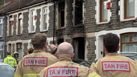 Fire fighters seen from behind in brown uniform with fire/tan written on the back, one drinking water from a bottle and looking at burnt out terraced house with blackened windows and brickwork