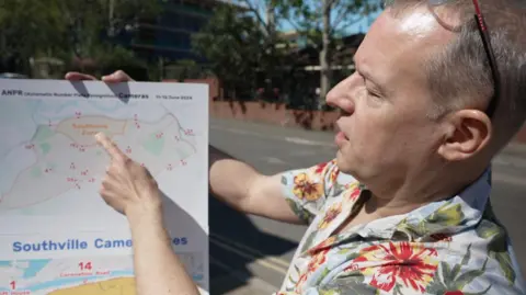 Matt is wearing a flowery shirt and has glasses on his head. He is pointing at a map he has printed out showing the camera positions that were used to collect data for the South Bristol Liveable Neighbourhood.