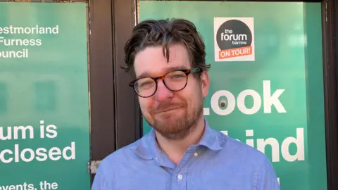 Walney Musical Theatre Society's chairman John Edwards. He has short wavy dark hair and blue eyes. He wears glasses and a blue shirt. He is looking at the camera with a slight smile. Behind him are posters on The Forum's windows saying the Barrow venue is closed. 