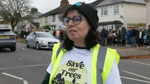 A woman wearing a t-shirt which says '"save our trees". She has a green high visibility jacket on and is also wearing a black woollen hat and glasses. 