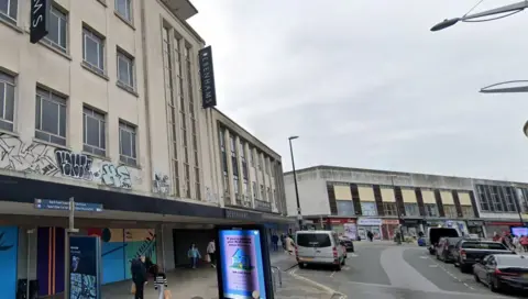 A shot of a shopping street. A Debenhams department store sign is visible but the shop is boarded up. Cars are parked on either side of the road. 