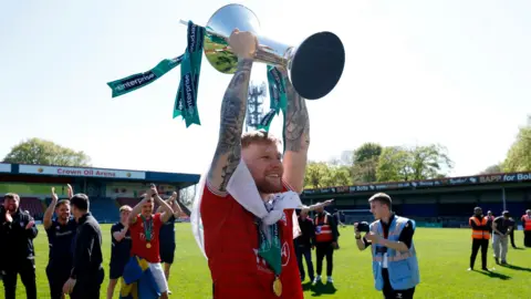 Cody Froggatt/PA Wire A bearded football player in red kit holds a silver trophy aloft and smiles. In the background, other players and club staff applaud and smile.
