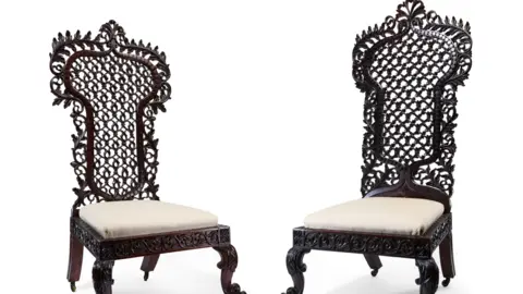Two chairs with tall backs that have been carved in an intricate style are pictured against a white backdrop. The chairs are made of a dark wood and have white cushions on them where a person would sit.